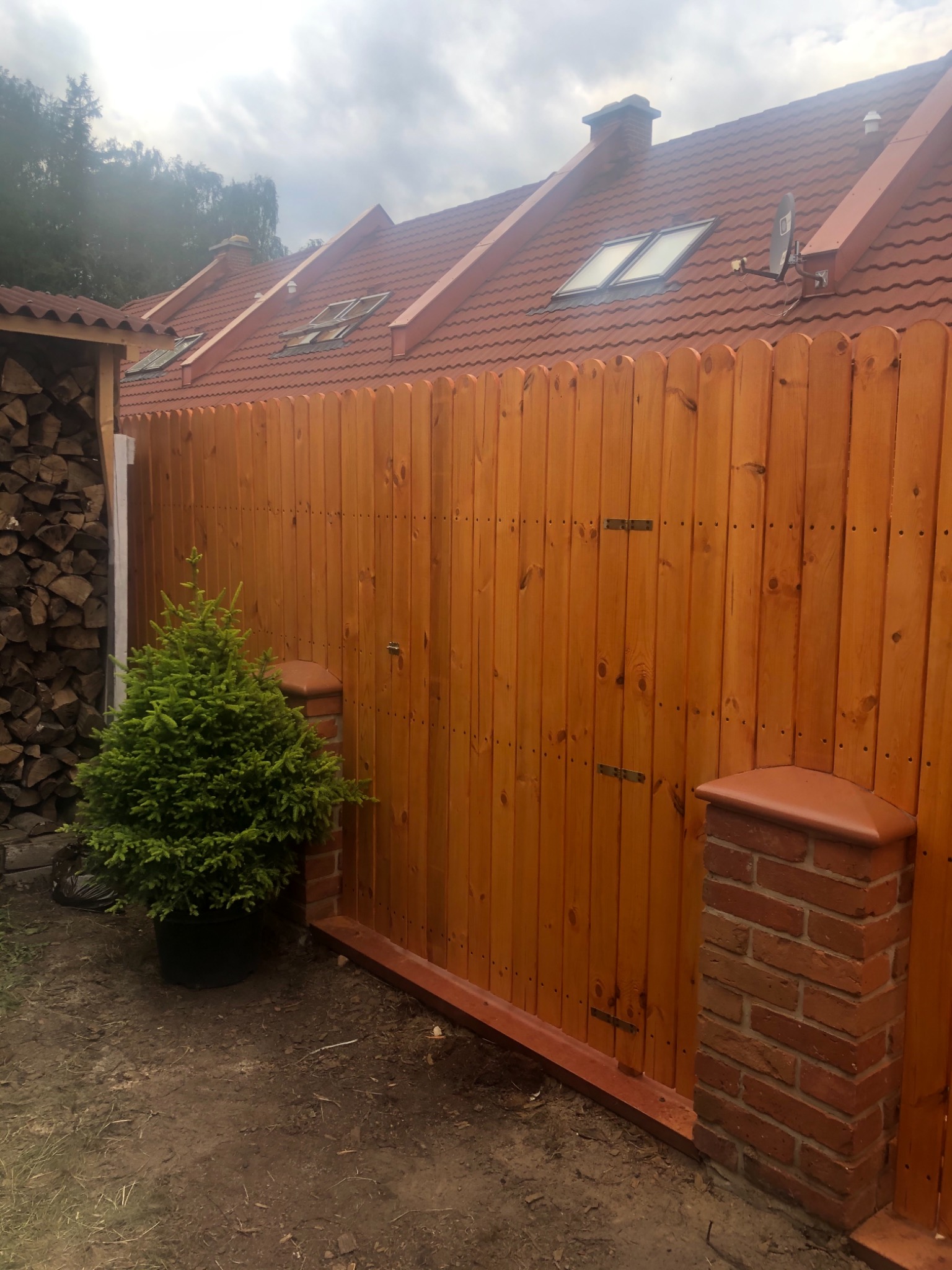 Nowo postawione drewniane ogrodzenie z bramą przesuwną i ceglanymi słupkami, widok na dom z czerwonym dachem i drzewa w tle, mała choinka w doniczce przy ogrodzeniu.