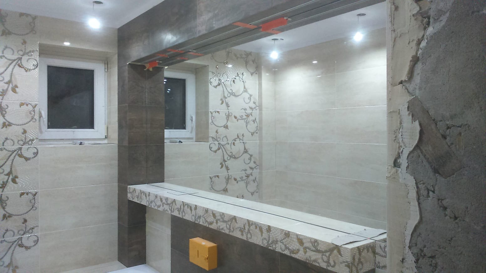 Łazienka w trakcie wykańczania: beżowe i brązowe płytki z motywem roślinnym, surowa ściana z resztkami tynku, widoczny stelaż pod sufitem.