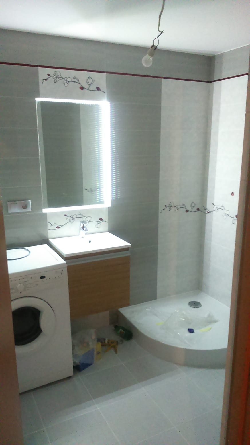 Wykończona łazienka z szarymi płytkami, kabiną prysznicową, umywalką z szafką i pralką; na ścianach dekoracyjny motyw roślinny, surowa żarówka w suficie.