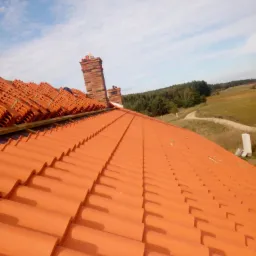 Pomarańczowa dachówka ceramiczna ułożona na nowym dachu, widoczne kominy z cegły i elementy mocujące dachówki przed ostatecznym montażem.