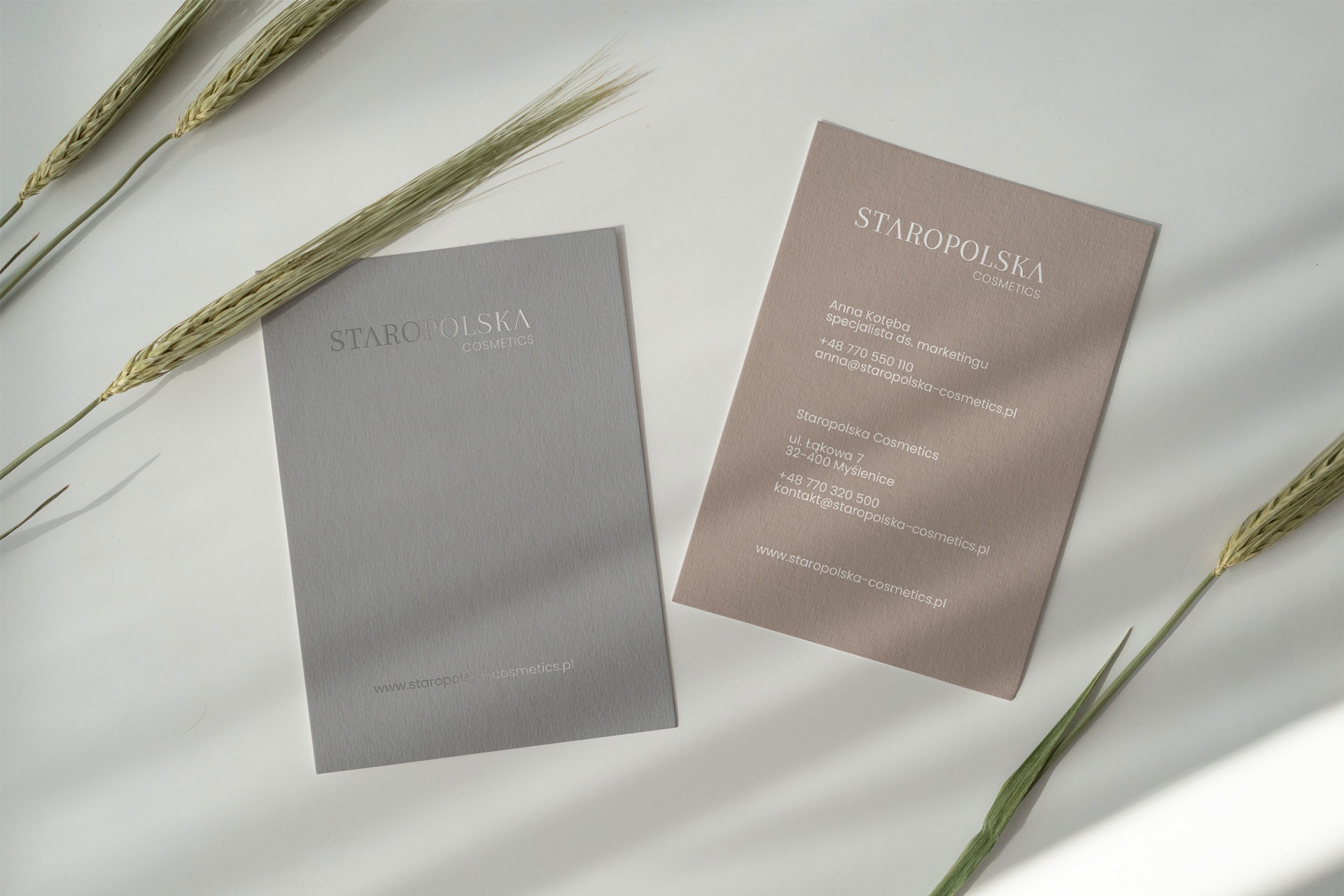 Dwie wizytówki Staropolska Cosmetics w odcieniach szarości i beżu, ułożone na białym tle z elementami roślinnymi, prezentujące minimalistyczny design i dane kontaktowe firmy.