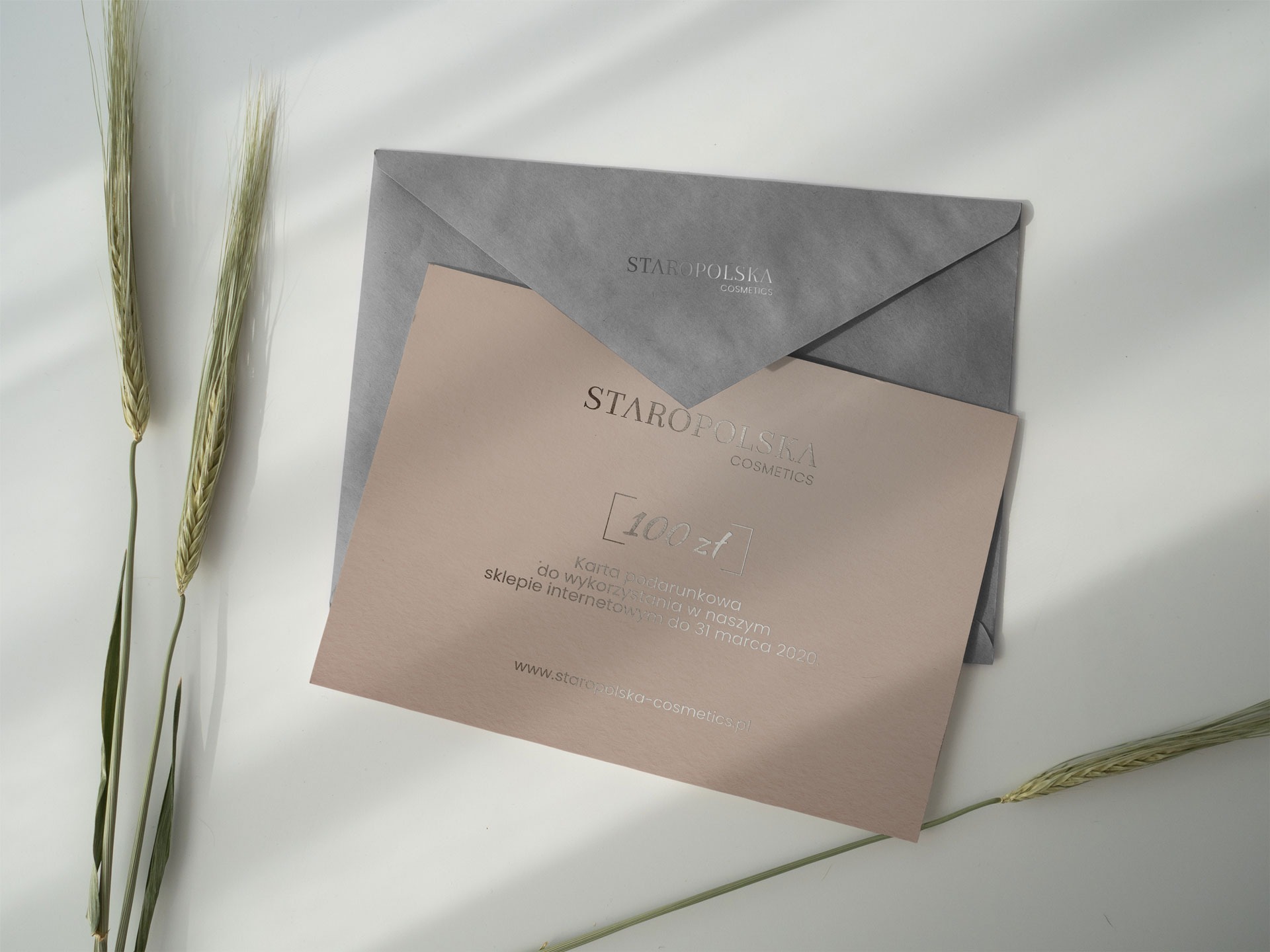 Karta podarunkowa o wartości 100 zł od Staropolska Cosmetics leżąca obok szarej koperty i dwóch źdźbeł zboża na białym tle, delikatne cienie