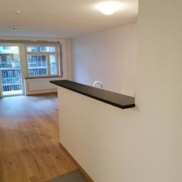 Yan Biryla - Wnętrze mieszkania po remoncie, widoczna drewniana podłoga, świeżo pomalowane ściany i fragment baru z czarnym blatem oraz widok na balkon.