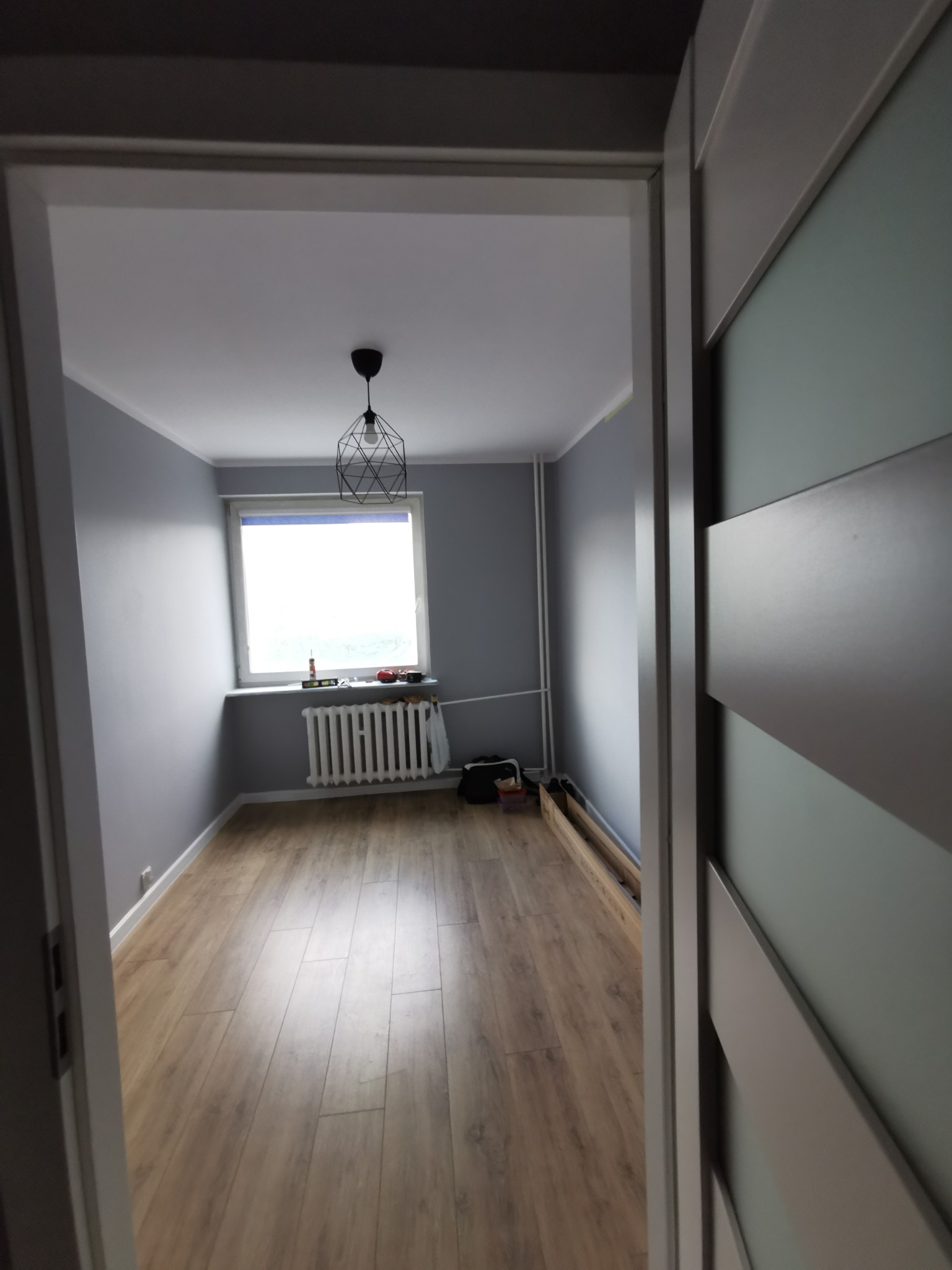 Pomalowane na szaro wnętrze pokoju z panelami podłogowymi, widok zza uchylonych drzwi, w oknie grzejnik i narzędzia.