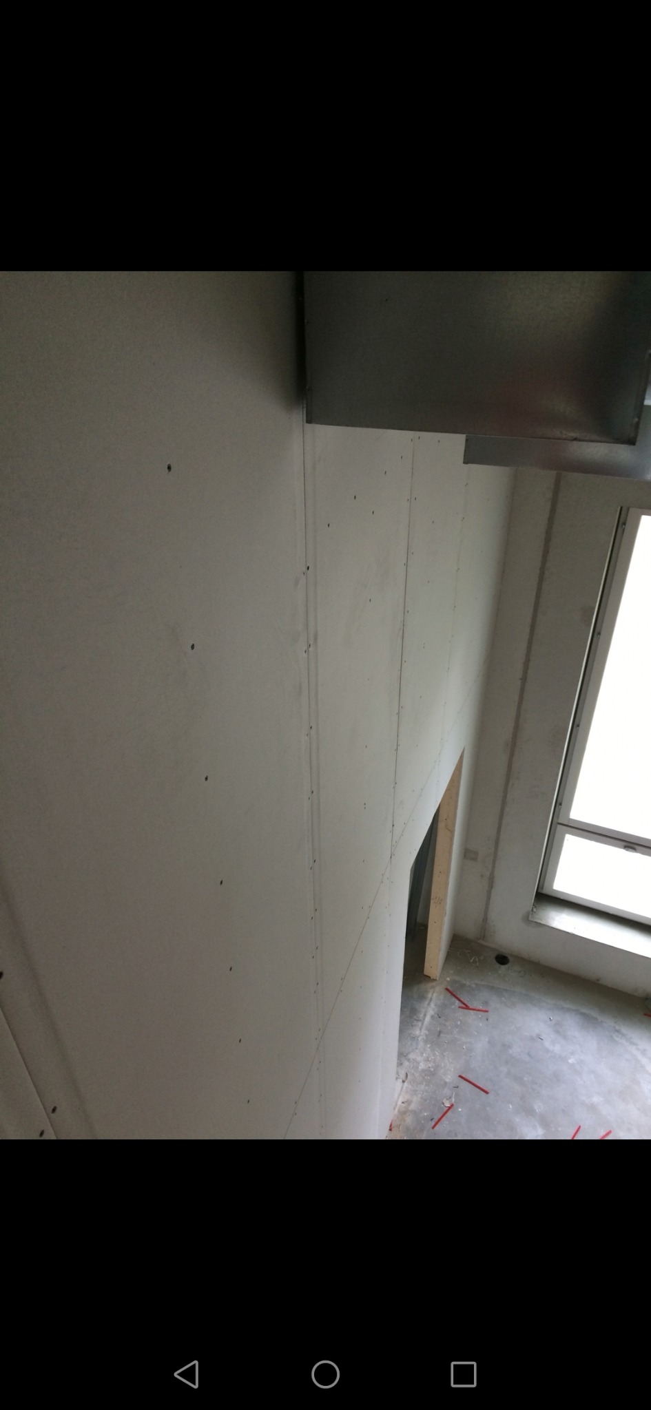 Wnętrze w trakcie prac wykończeniowych; ściany pokryte płytami, widoczne łączenia i otwór drzwiowy, podłoga z resztkami po budowie.