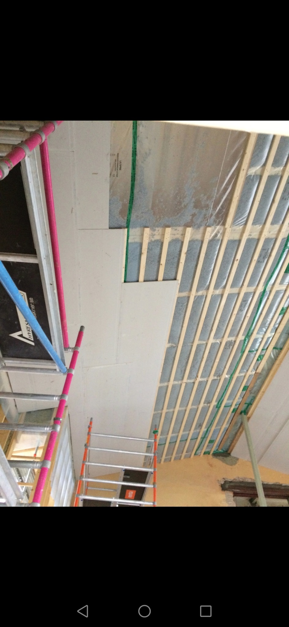 Montaż na stelażu sufitowym płyt. Widoczne profile, izolacja, fragment rusztowania z różowymi elementami.
