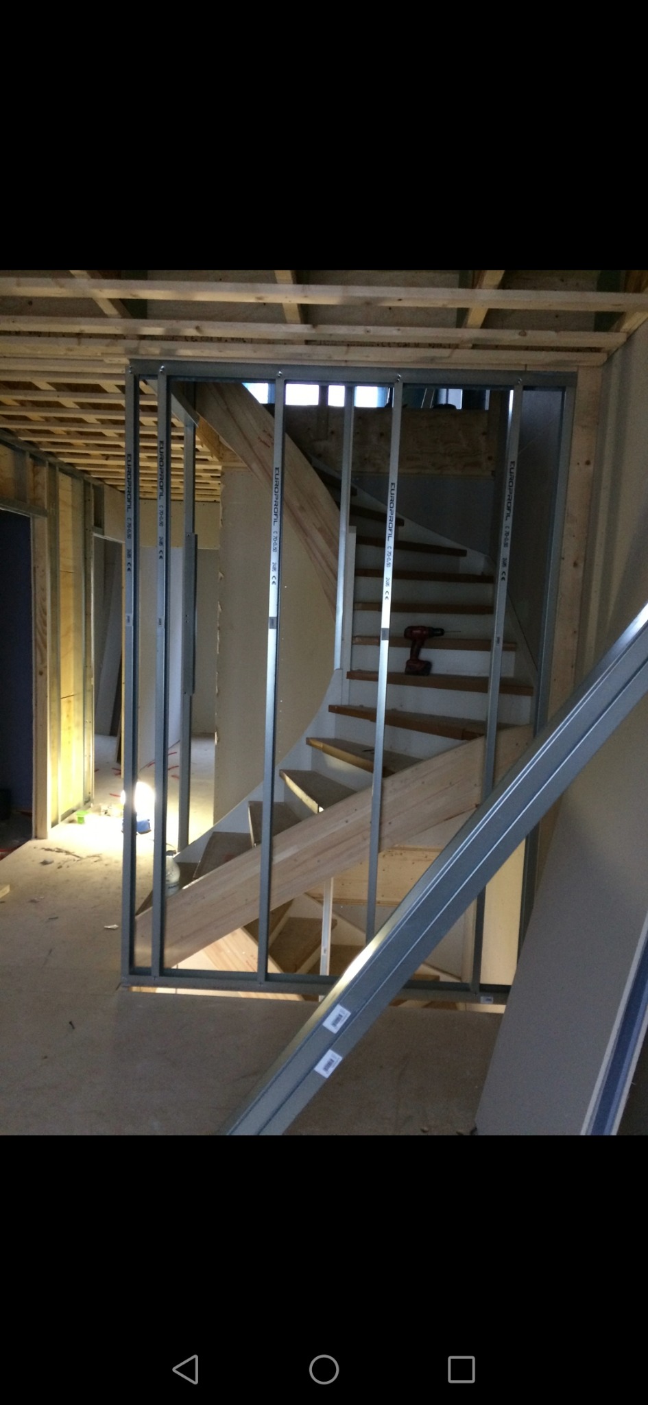 Wnętrze w trakcie remontu: metalowa konstrukcja ścianki działowej, widoczne drewniane schody i sufit, w tle narzędzie akumulatorowe na schodach.