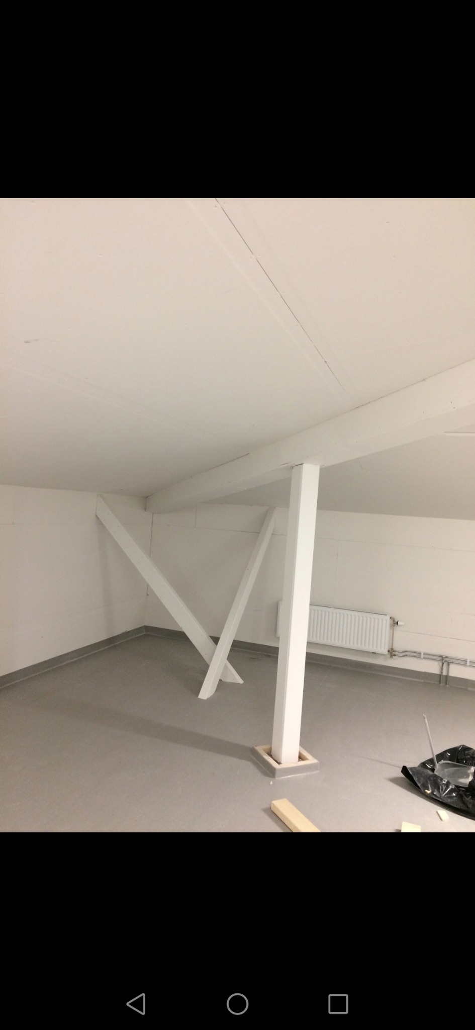 Wnętrze pomieszczenia z białymi ścianami, szarą podłogą i wzmocnieniem stropu w postaci białych, drewnianych belek i słupa, widoczny grzejnik na ścianie.
