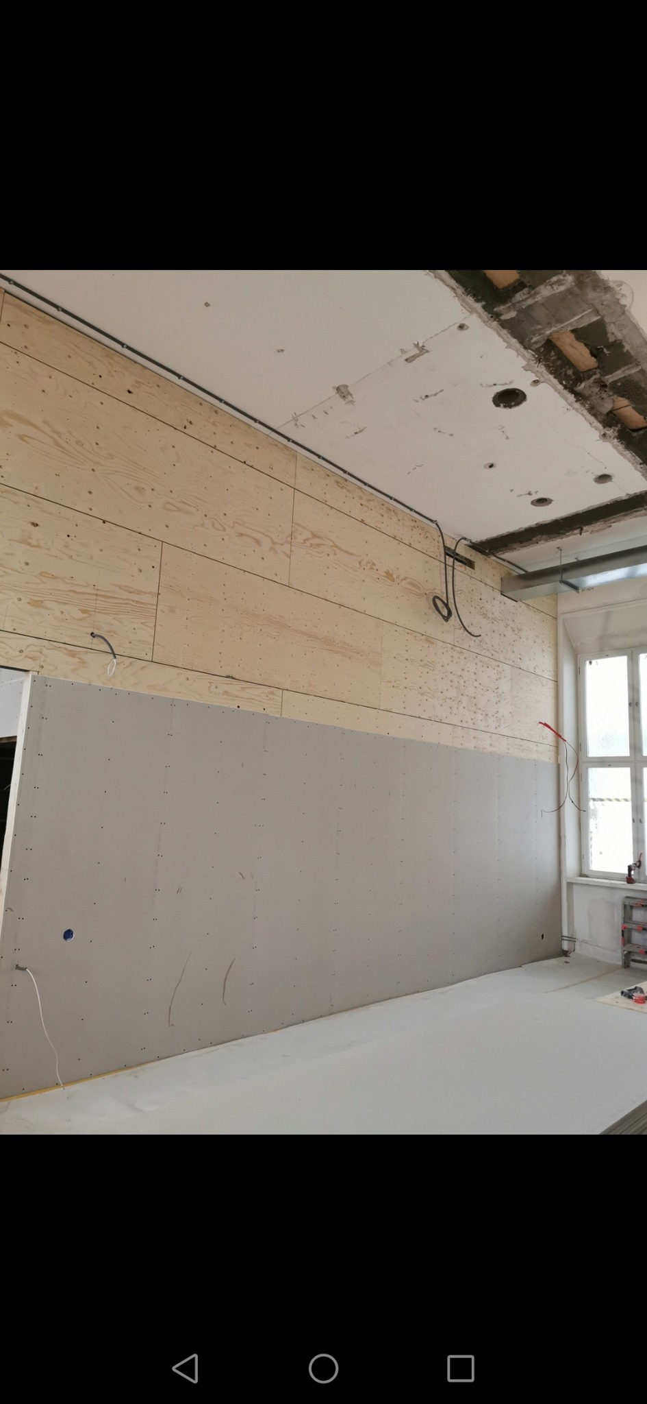 Wnętrze w trakcie prac wykończeniowych: ściana z drewnianych paneli w górnej części, poniżej zamontowana płyta gipsowo-kartonowa, widoczne kable i surowy sufit.