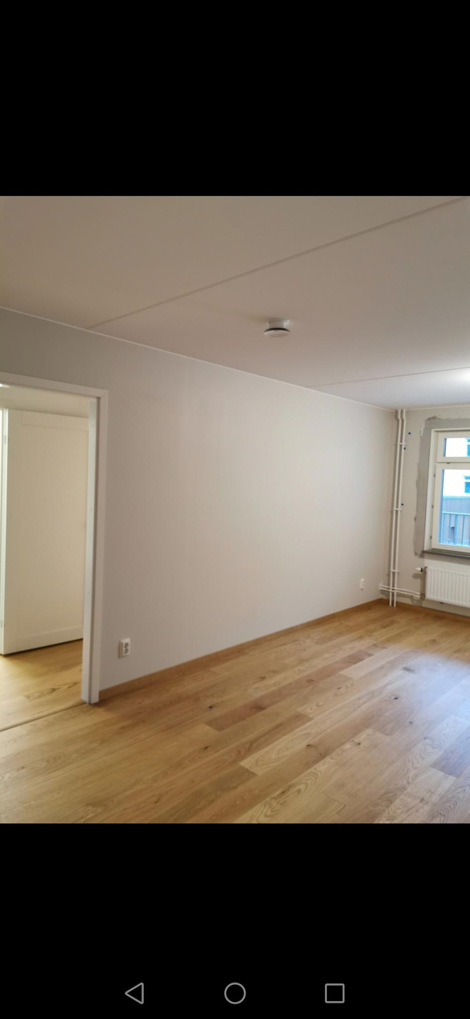 Jasny, pomalowany pokój z drewnianą podłogą, białymi listwami przypodłogowymi i widocznym czujnikiem dymu na suficie. Okno z grzejnikiem i rurami grzewczymi po prawej stronie. Otwór drzwiowy...