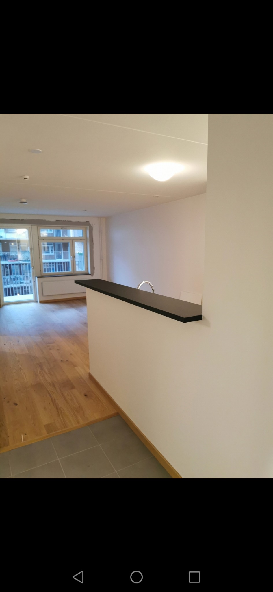 Wnętrze mieszkania po remoncie, widoczna drewniana podłoga, świeżo pomalowane ściany i fragment baru z czarnym blatem oraz widok na balkon.