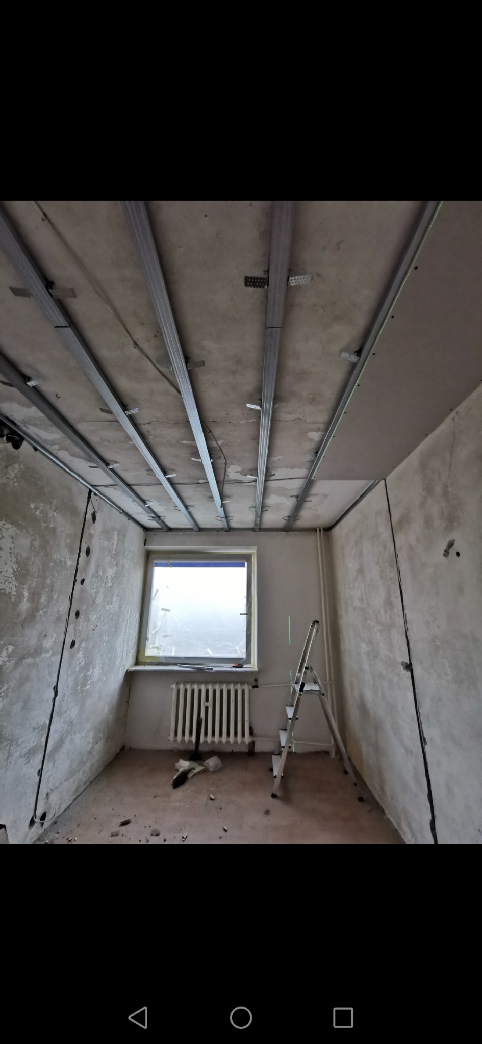 Metalowa konstrukcja sufitowa z zamocowanymi wieszakami, przygotowana pod montaż płyt w remontowanym pokoju z oknem i drabinką.