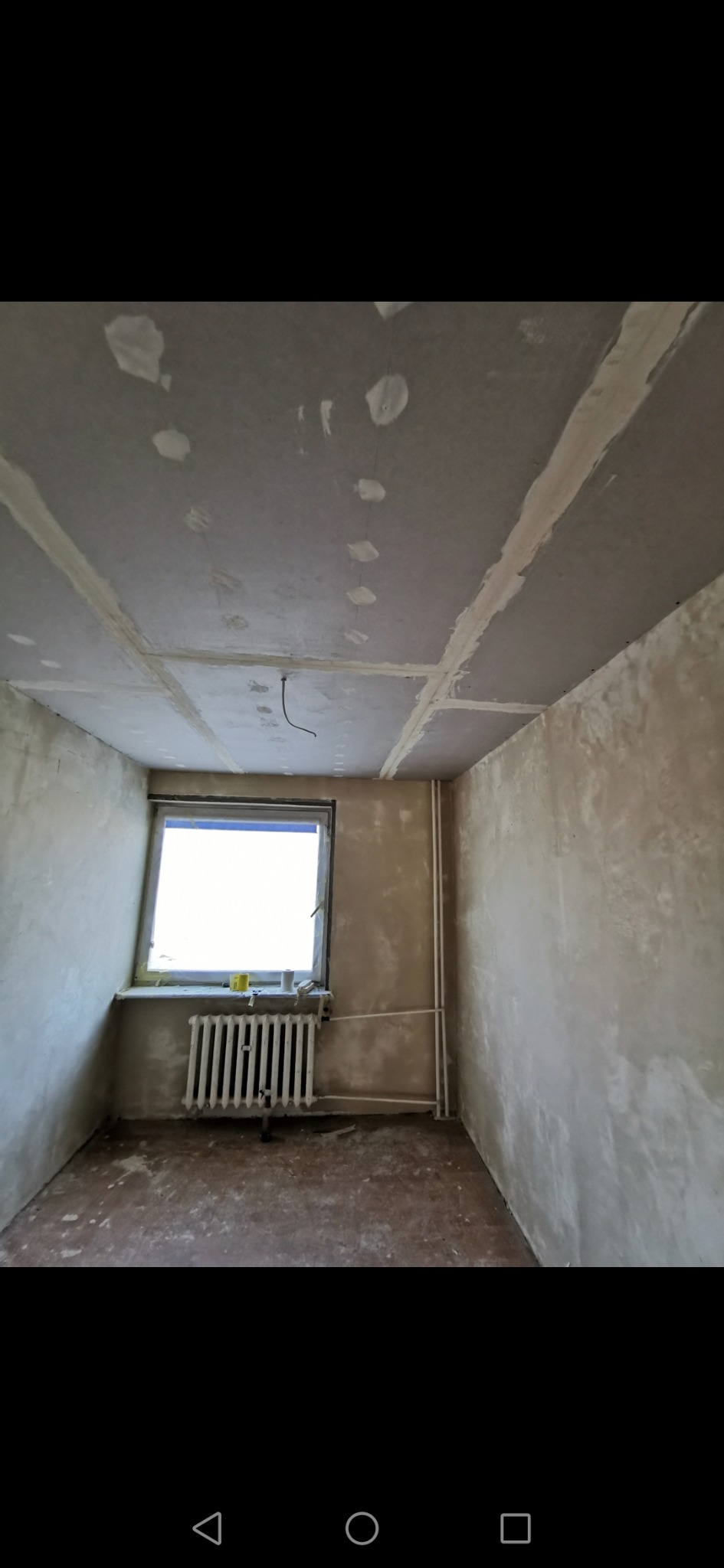 Pokój w trakcie remontu: surowy sufit z widocznymi łączeniami płyt, ściany w szpachli, grzejnik pod oknem, widoczne instalacje grzewcze.