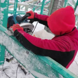 Pracownik w czerwonej kurtce z kapturem, w trakcie cięcia drewnianej belki piłą tarczową na budowie dachu pokrytej śniegiem.