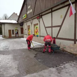 Dwóch robotników w czerwonych kombinezonach układa kostkę brukową na oblodzonym placu przed sklepem spożywczym z drewnianą elewacją, widoczna taczka i flaga Polski.