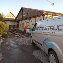 Przygotowanie podłoża pod fundament budynku sklepu spożywczego, widoczny pracownik w czerwonym kombinezonie z taczką oraz srebrny samochód dostawczy z logo firmy budowlanej.