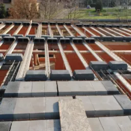 Wieniec stropowy na murze z pustaków ceramicznych, widoczne zbrojenie i betonowe nadproża, w tle zabudowa jednorodzinna.