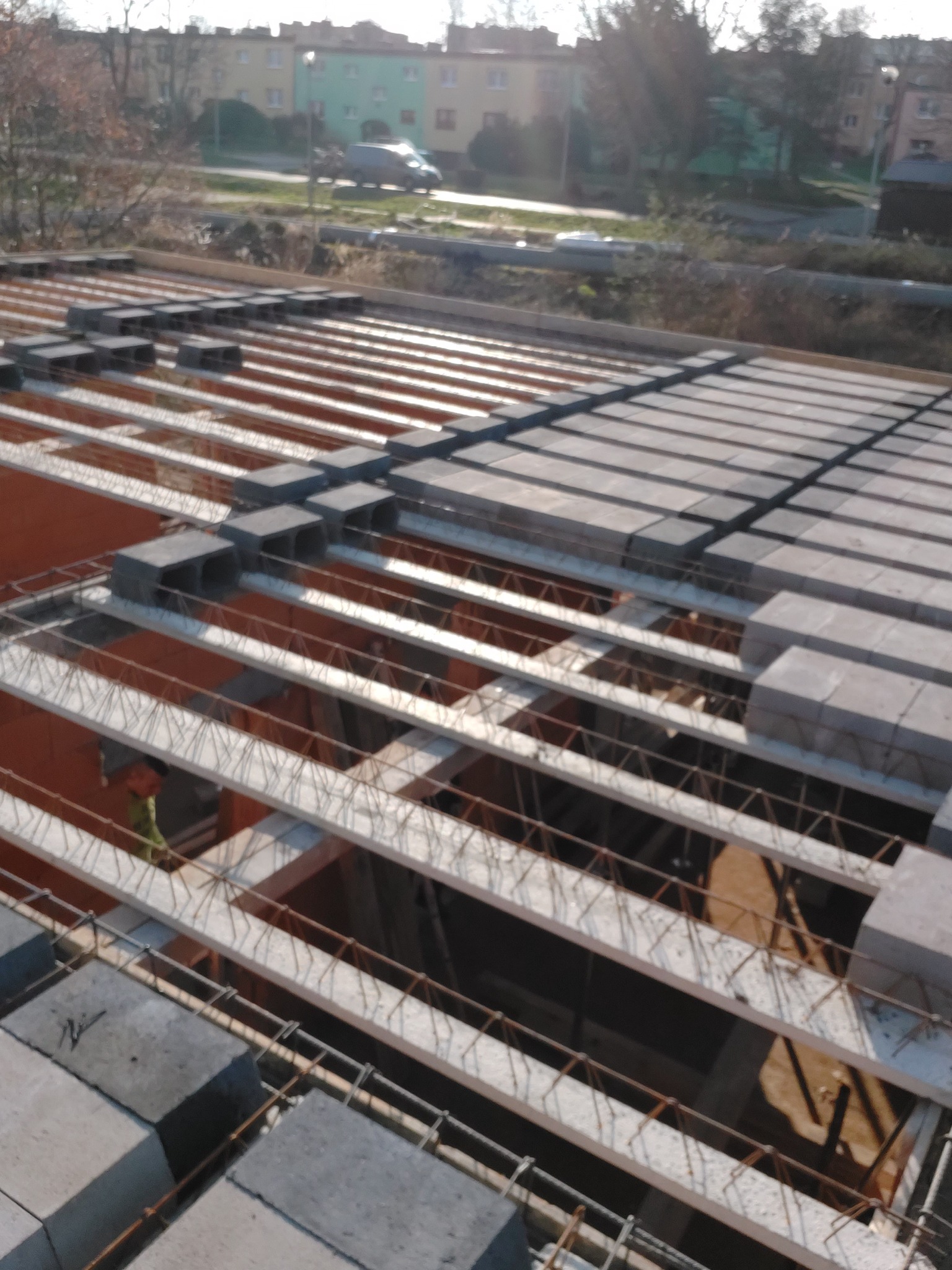 Widok z góry na strop Teriva w trakcie montażu, z widocznymi belkami, pustakami betonowymi oraz zbrojeniem, w tle budynki mieszkalne i drzewa.