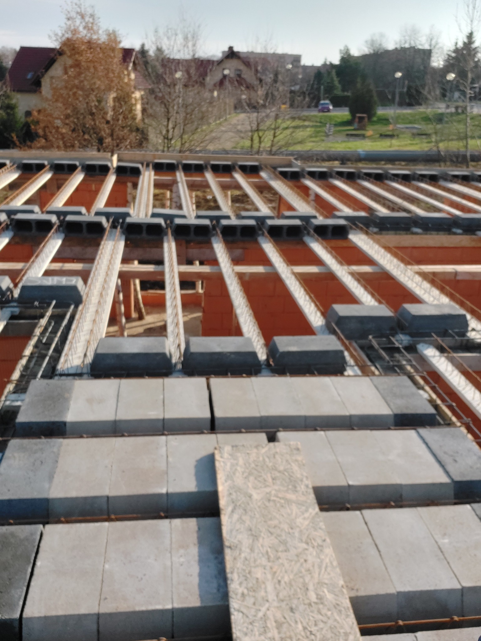 Wieniec stropowy na murze z pustaków ceramicznych, widoczne zbrojenie i betonowe nadproża, w tle zabudowa jednorodzinna.