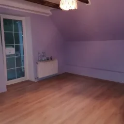 Puste poddasze z drewnianą podłogą, świeżo pomalowanymi fioletowymi ścianami, grzejnikiem pod oknem i drewnianymi belkami stropowymi.