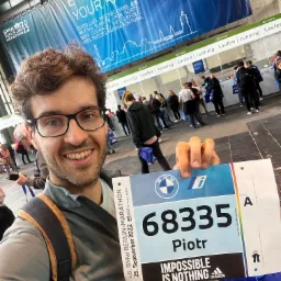 Uśmiechnięty mężczyzna z okularami trzyma numer startowy 68335 Piotra na 48. BMW Berlin-Marathon 2022, w tle widoczny tłum i baner z napisem 'Enjoy Your Race'.
