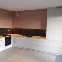 D&S meble na wymiar Sebastian Durlik - Nowoczesna kuchnia w odcieniach bieli i drewna, z czarnym panelem nad blatem, minimalistyczne fronty szafek, widoczne niedokończone instalacje elektryczne na suficie.