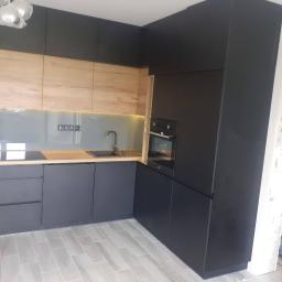 D&S meble na wymiar Sebastian Durlik - Nowoczesna kuchnia narożna z czarnymi i drewnopodobnymi frontami, szarym szkłem nad blatem i płytą indukcyjną, widok na zlewozmywak i piekarnik w zabudowie, szare płytki podłogowe, fragment...