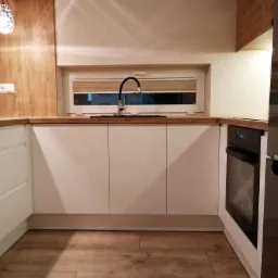 Nowoczesna kuchnia z białymi frontami szafek, drewnianym blatem i podłogą, czarną armaturą przy oknie z żaluzjami, oraz częściowo widoczną zabudowaną lodówką.