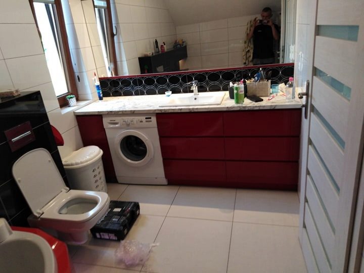 Czerwona zabudowa szafkowa pod umywalką z pralką w małej łazience na poddaszu, widok na lustro z odbiciem fotografa.