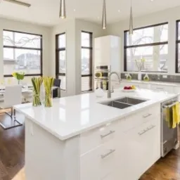 Jasna kuchnia z białymi szafkami i wyspą z błyszczącym blatem, widok na stół jadalny i duże okna.