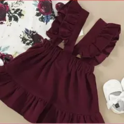 Komplet niemowlęcy: bordowa sukienka na szelkach z falbankami i bluzka w kwiaty, obok białe buciki.