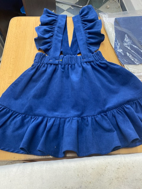 Niebieska sukienka dziecięca z falbankami, uszyta z prążkowanego materiału, z widocznymi szpilkami przy gumce w pasie, leżąca na stole.