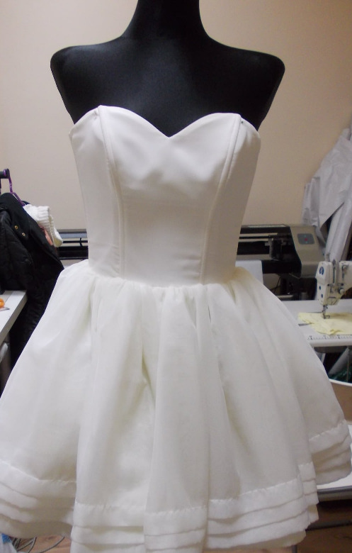 Biała, krótka sukienka z tiulowym dołem na czarnym manekinie krawieckim, widoczna maszyna do szycia w tle.