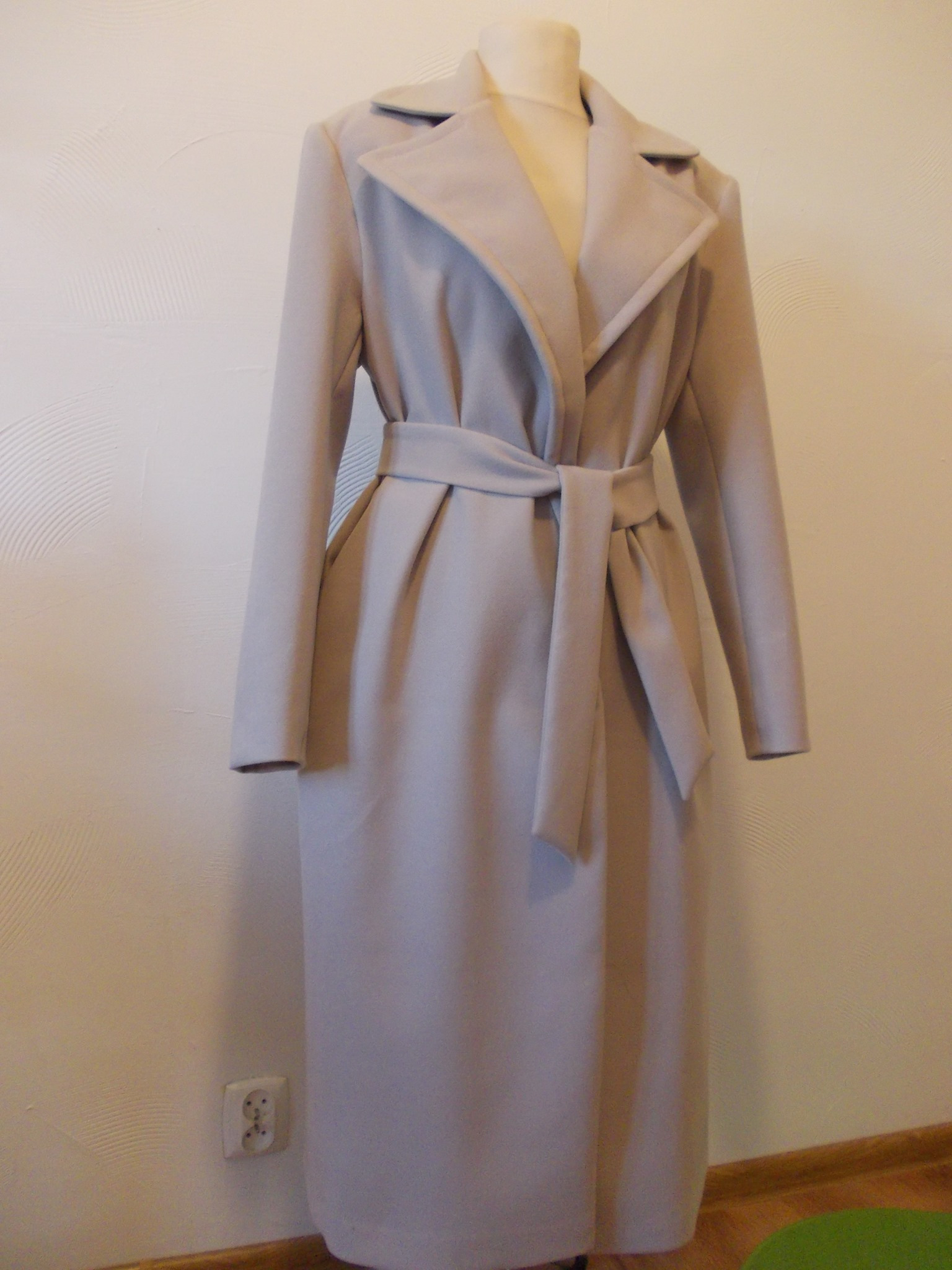 Elegancki, długi płaszcz damski w odcieniu jasnego beżu, zapinany na wiązany pasek, prezentowany na manekinie na tle ściany z widocznym gniazdkiem elektrycznym.