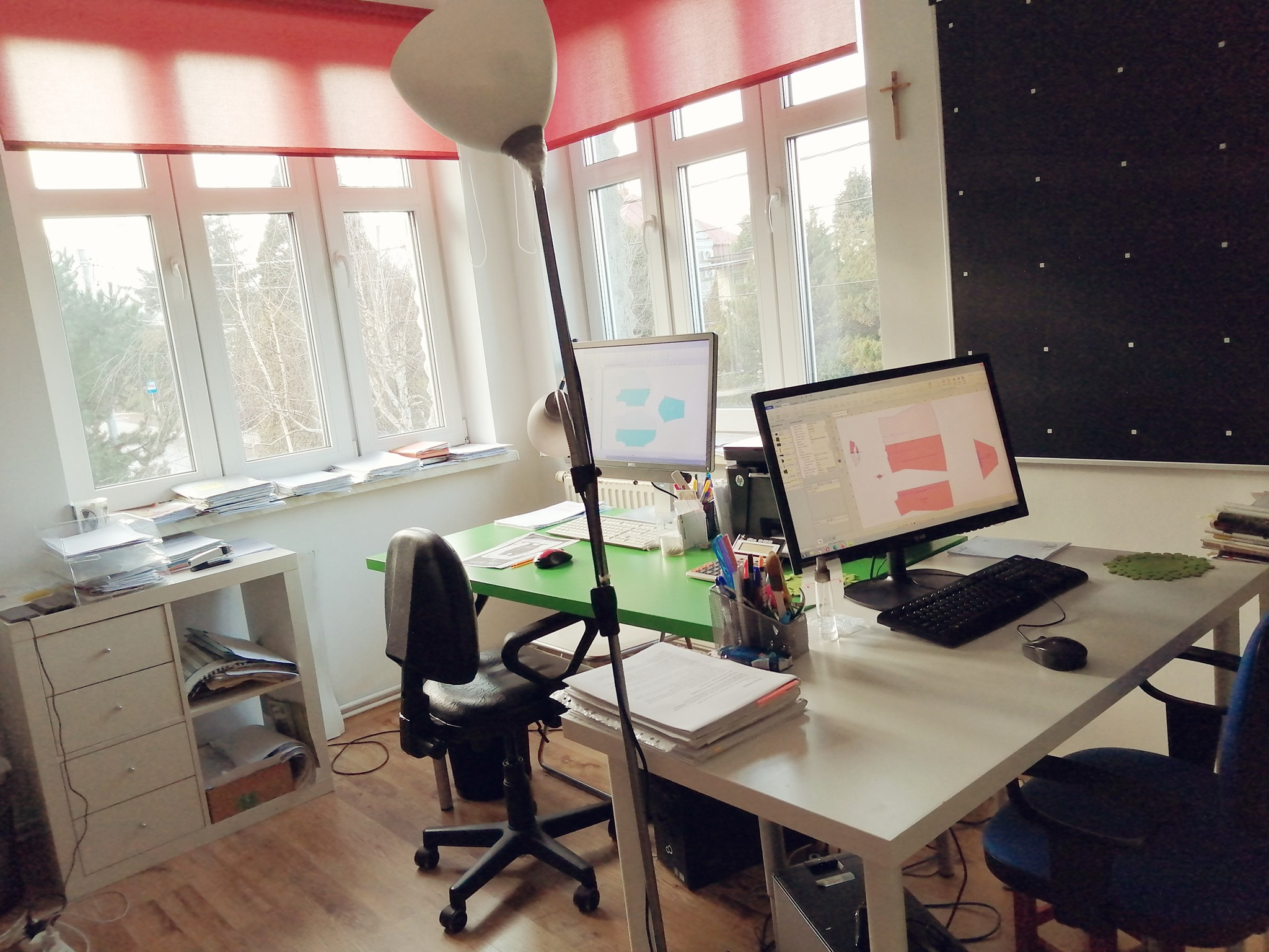 Stanowisko pracy z dwoma monitorami prezentującymi projekty odzieży, biurko z zielonym blatem, lampa stojąca, okno z roletami i czarna tablica z białymi punktami w tle.