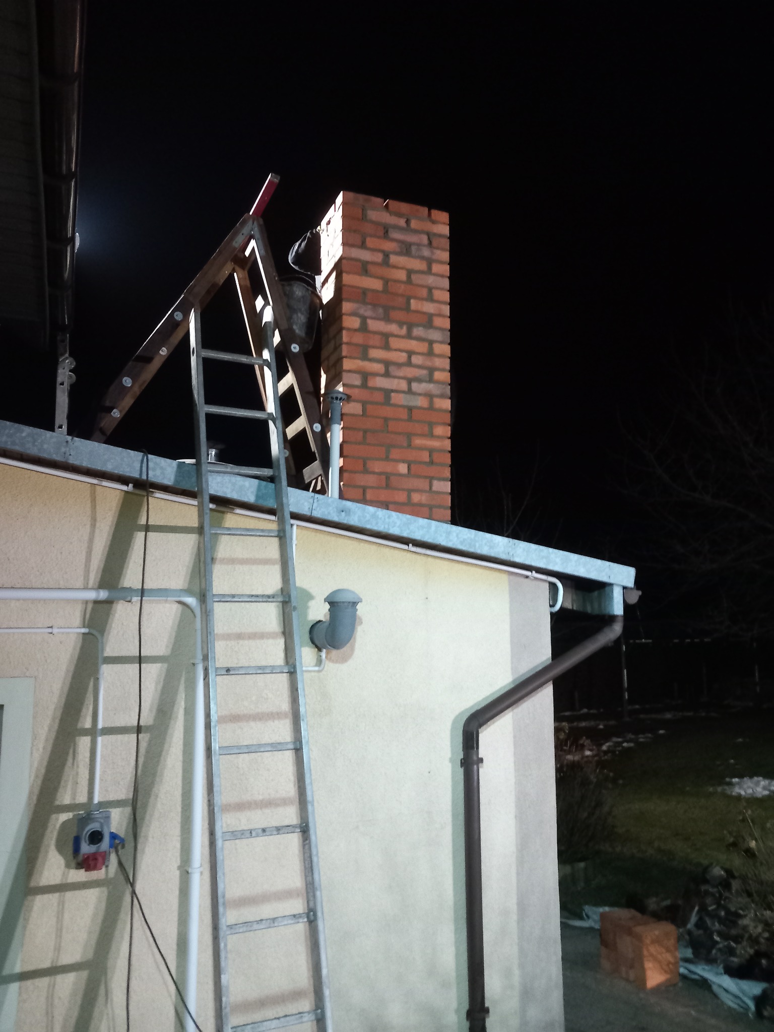 Metalowa drabina oparta o ścianę domu, prowadząca do ceglanego komina na dachu, nocne oświetlenie.