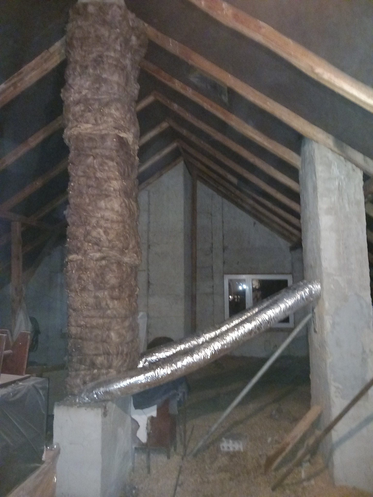 Ocieplony przewód kominowy na poddaszu, widoczne drewniane elementy konstrukcji dachu i przewód wentylacyjny.