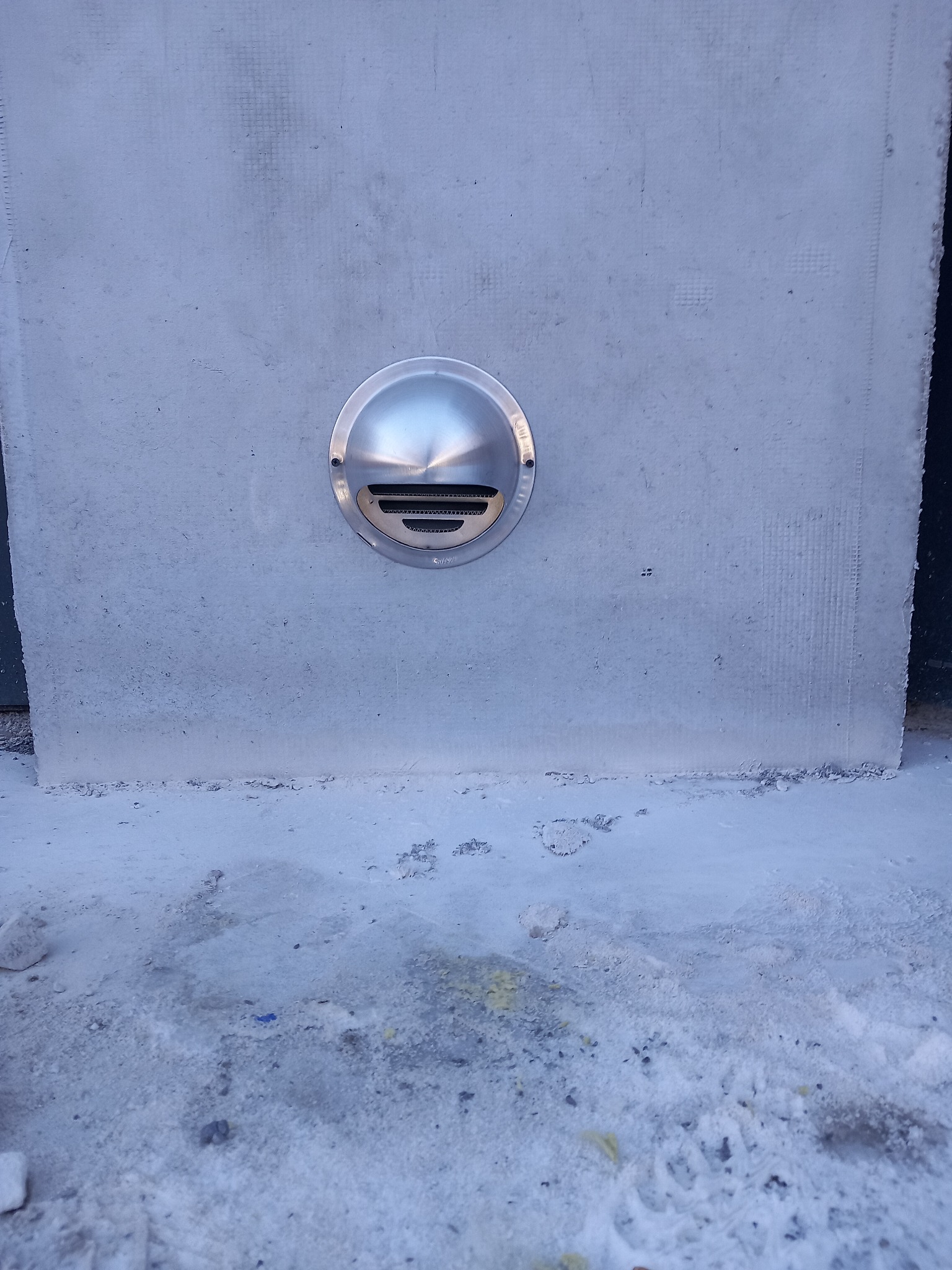 Okrągła, metalowa kratka wentylacyjna zamontowana na szarej, betonowej ścianie, z widocznymi śladami zaprawy na podłożu.