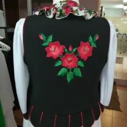 Czarna kamizelka ludowa z haftowanymi czerwonymi różami i zielonymi liśćmi, wykończona czerwoną lamówką, na manekinie z białą bluzką i kwiecistą falbaną przy szyi.