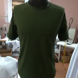 Zielona koszulka z krótkim rękawem na manekinie w pracowni krawieckiej, widoczne maszyny do szycia w tle.