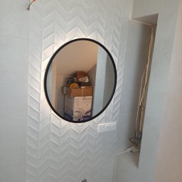 Marcin Bielecki - Biała łazienka z wzorzystymi płytkami, okrągłe lustro z podświetleniem odbija kartony na poddaszu, widoczne dwa krany i przełącznik światła.