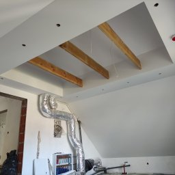 Marcin Bielecki - Wnętrze poddasza w trakcie prac wykończeniowych: widoczne drewniane belki w suficie, instalacja wentylacyjna w srebrnej folii, ściany przygotowane pod malowanie, otwór drzwiowy w stanie surowym.