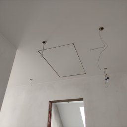 Marcin Bielecki - Białe ściany i sufit w trakcie prac wykończeniowych, widoczne otwory na oświetlenie i rama otworu rewizyjnego w suficie, zwisające kable elektryczne, surowy otwór drzwiowy w tle.