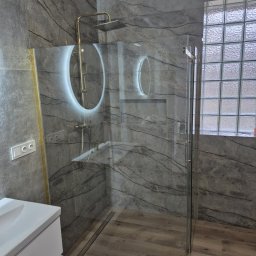 Marcin Bielecki - Nowoczesna łazienka z szarymi płytkami imitującymi marmur, kabiną prysznicową ze szkła hartowanego, złotą armaturą i okrągłym lustrem z podświetleniem LED.