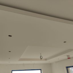 Marcin Bielecki - Wnętrze w Wołominie w trakcie prac wykończeniowych: widok na sufit podwieszany z wyciętymi otworami na oświetlenie, widoczne kable elektryczne i elementy konstrukcyjne.