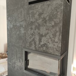 Marcin Bielecki - Szary kominek obłożony dekoracyjną strukturą tynku, z metalowymi elementami wykończeniowymi i czarnymi otworami wentylacyjnymi, widoczny fragment marmurowej podłogi i zabezpieczonej podłogi.