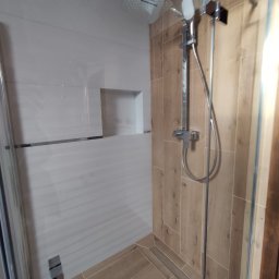 Marcin Bielecki - Nowoczesna kabina prysznicowa z deszczownicą i panelami ściennymi imitującymi drewno, widok z boku, minimalistyczny design z wnęką w ścianie wyłożonej białymi płytkami.