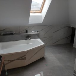 Marcin Bielecki - Jasna łazienka na poddaszu z oknem dachowym, wykończona płytkami imitującymi marmur z szarymi żyłami, z narożną wanną obudowaną tymi samymi płytkami i szarą półką nad wanną.