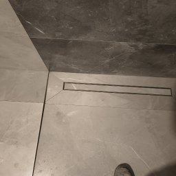 Marcin Bielecki - Szare płytki podłogowe i ścienne w łazience z odpływem liniowym, widoczny fragment buta.