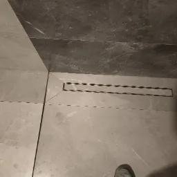 Szare płytki podłogowe i ścienne w łazience z odpływem liniowym, widoczny fragment buta.